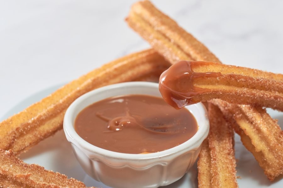 Traditional churros with sugar and cinnamon served on a marble table, with a bowl of caramel.